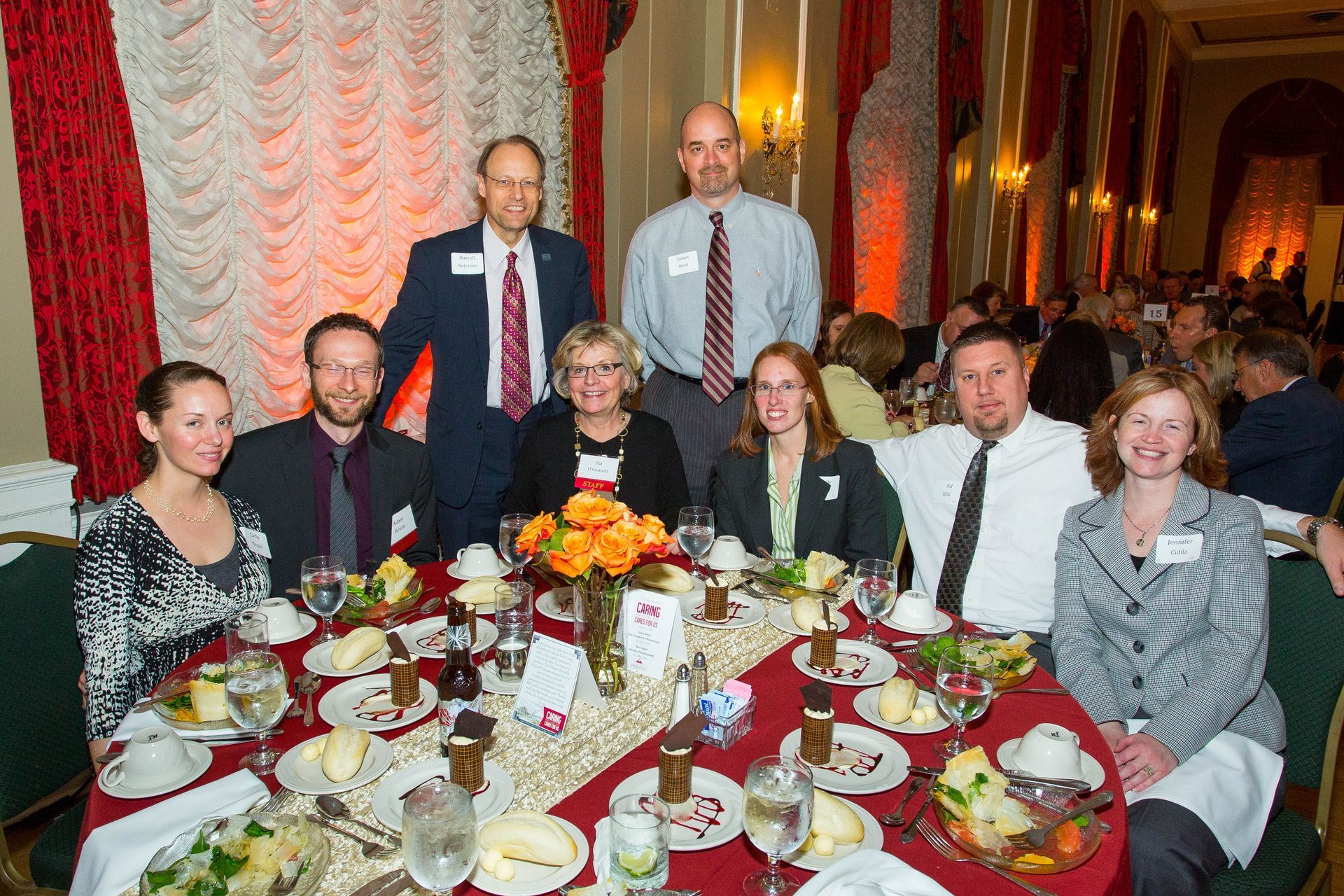 Group of people seated and standing around a formal banquet table with floral centerpieces and wine glasses