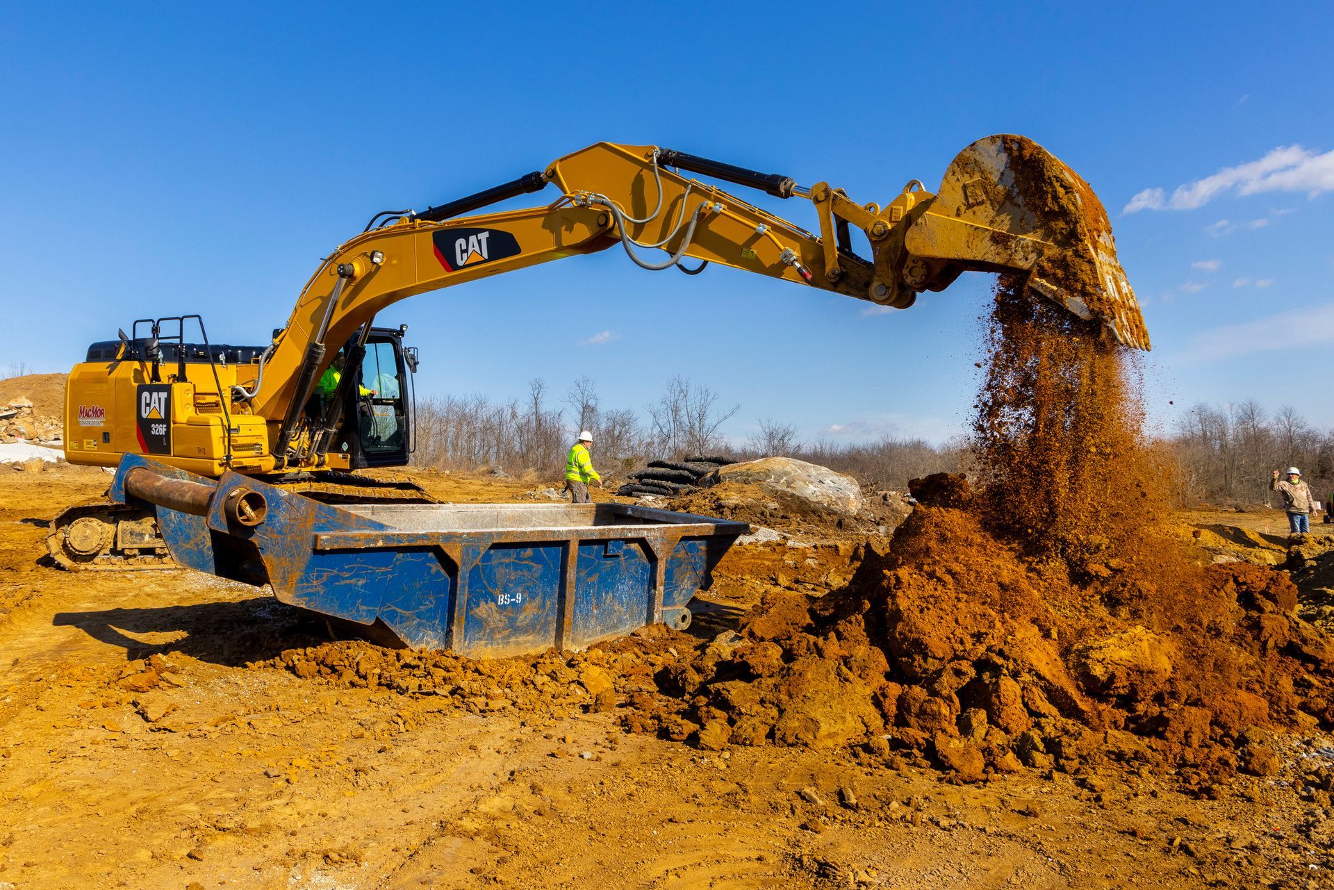 Yellow excavator dumping dirt into a blue truck at a muddy construction site under a clear sky