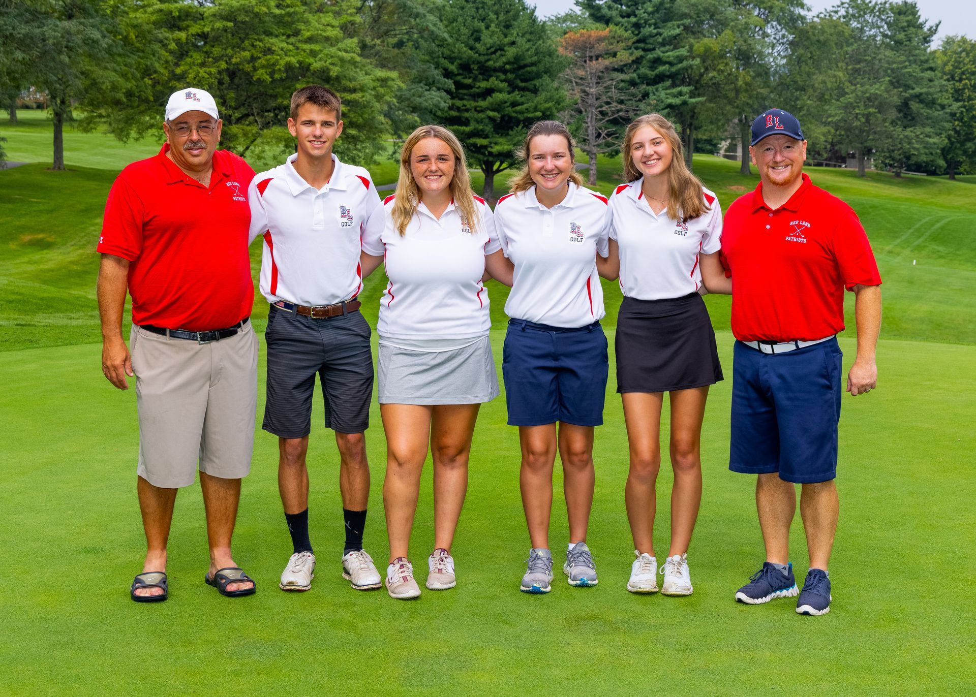 Six people in golf attire standing together on a golf course, smiling at the camera.
