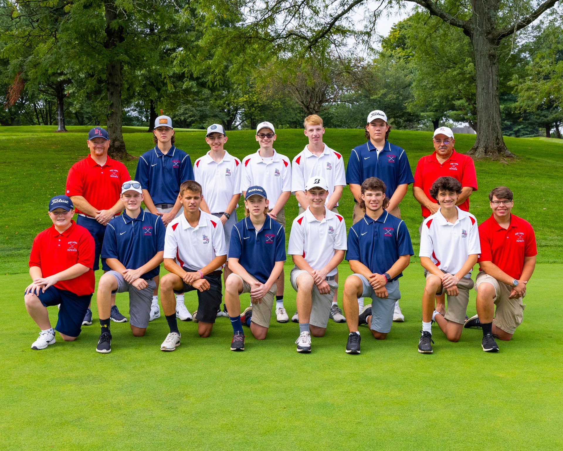 Group of golfers posing outdoors on a green lawn, wearing red, white, and blue team shirts.