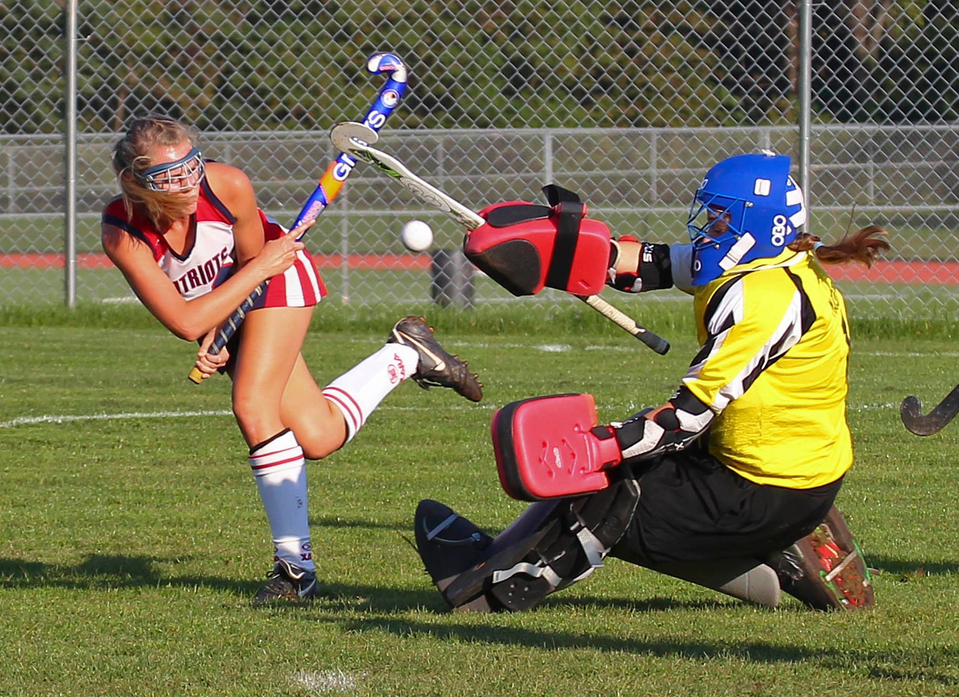 Field hockey player in red striking a ball while goalkeeper in yellow blocks the shot on grass field