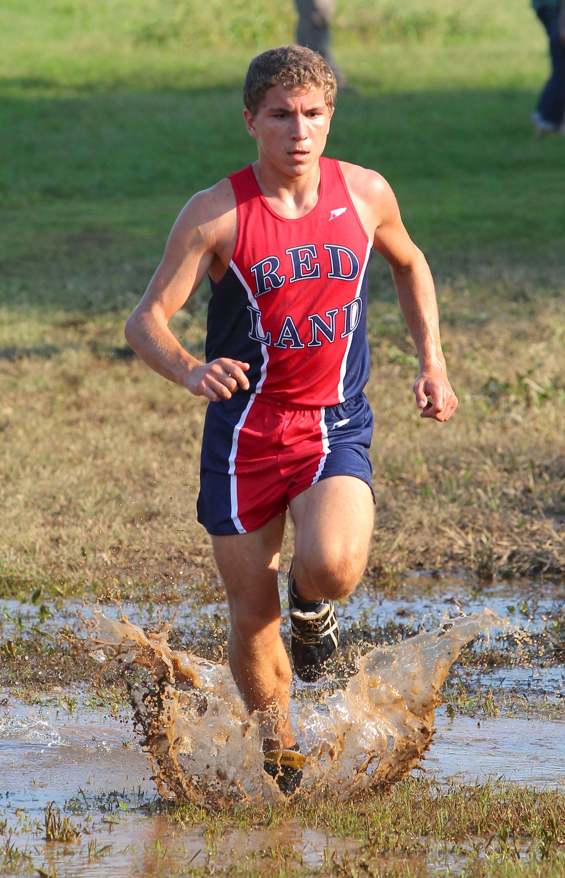Runner in red and blue uniform splashes through a muddy puddle on a grassy field.