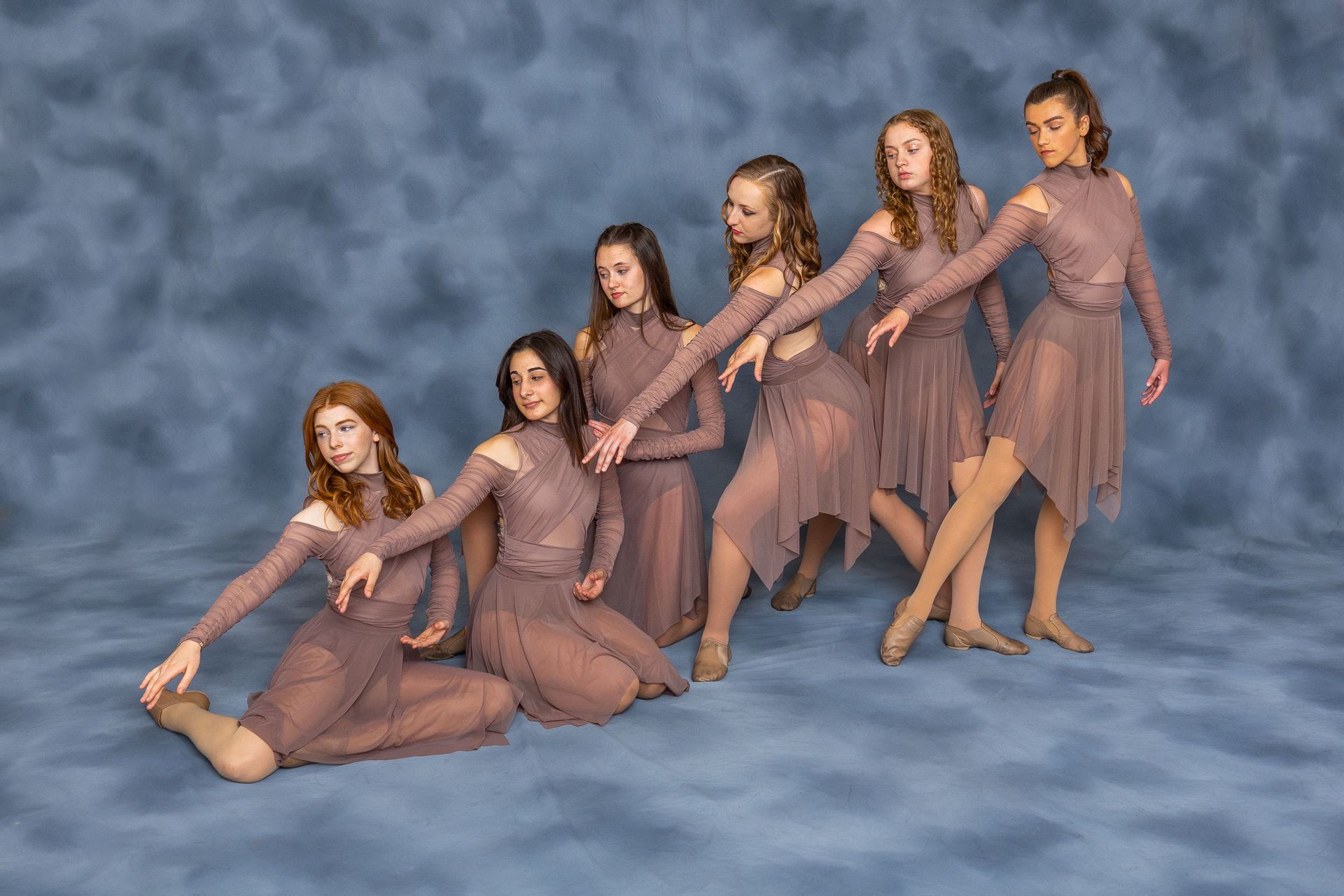 Six dancers in sheer pink costumes pose together on a blue studio backdrop.