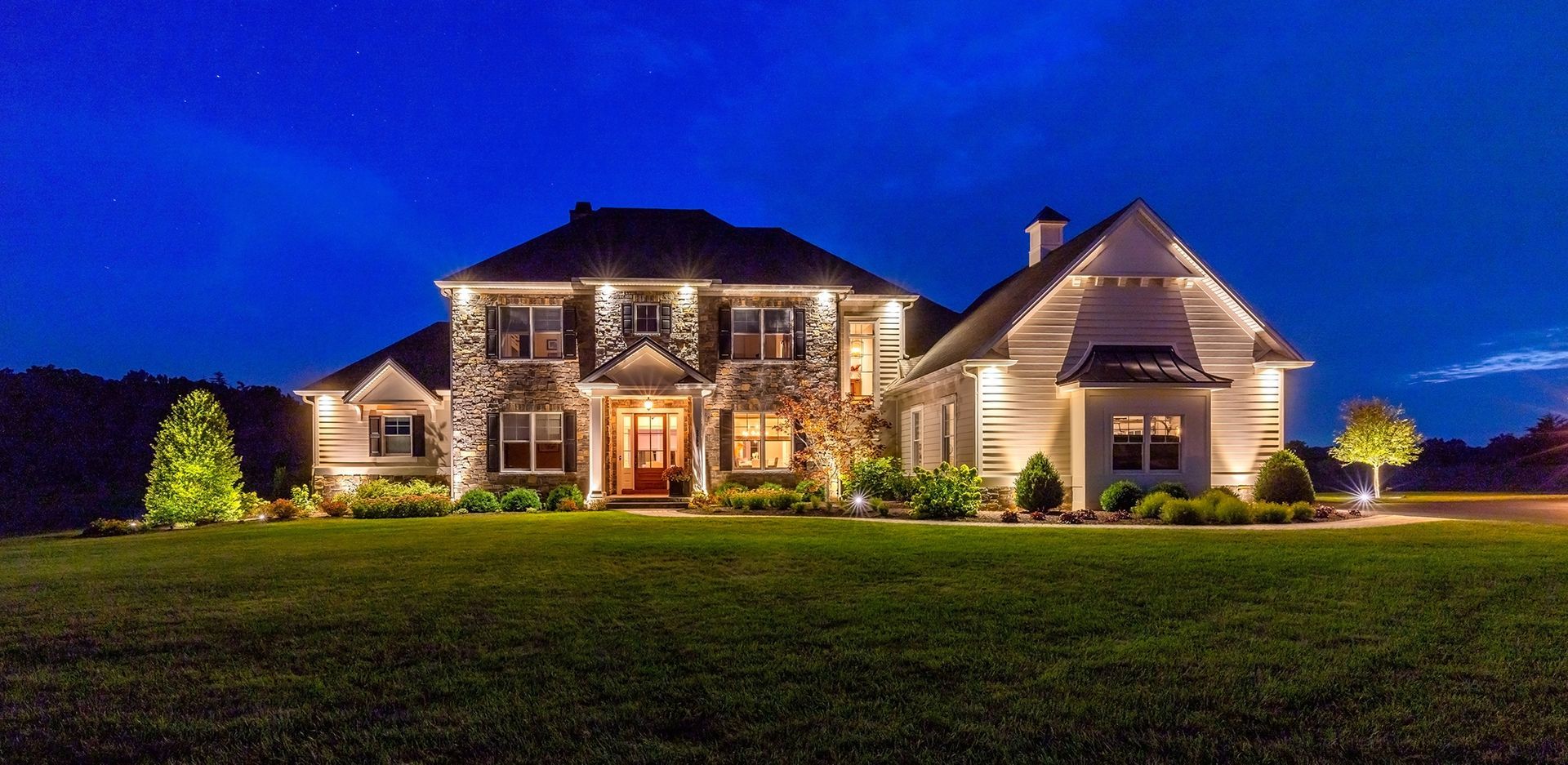 Illuminated stone house at dusk with a large lawn and warm exterior lights
