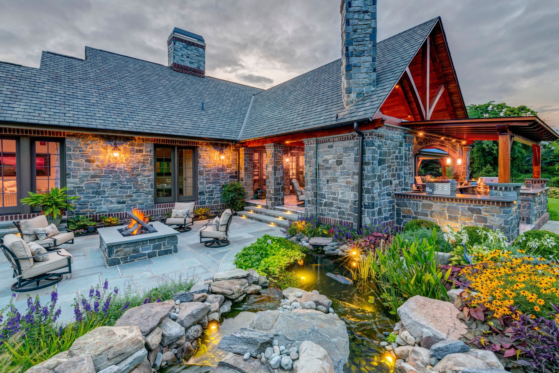 Stone house with a steep roof, lit patio seating, and a landscaped garden at dusk