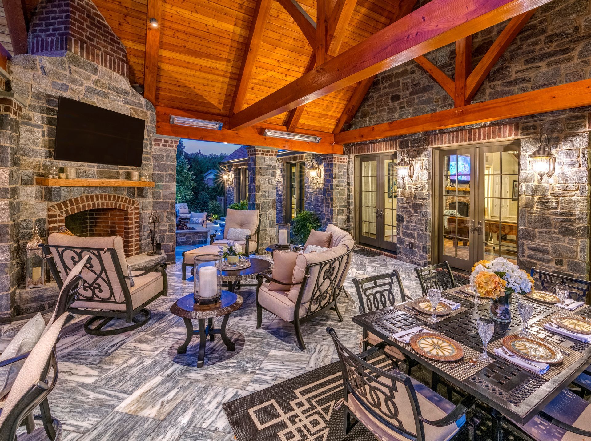 Cozy stone patio with a fireplace, wooden beams, cushioned chairs, and a dining table under warm lighting