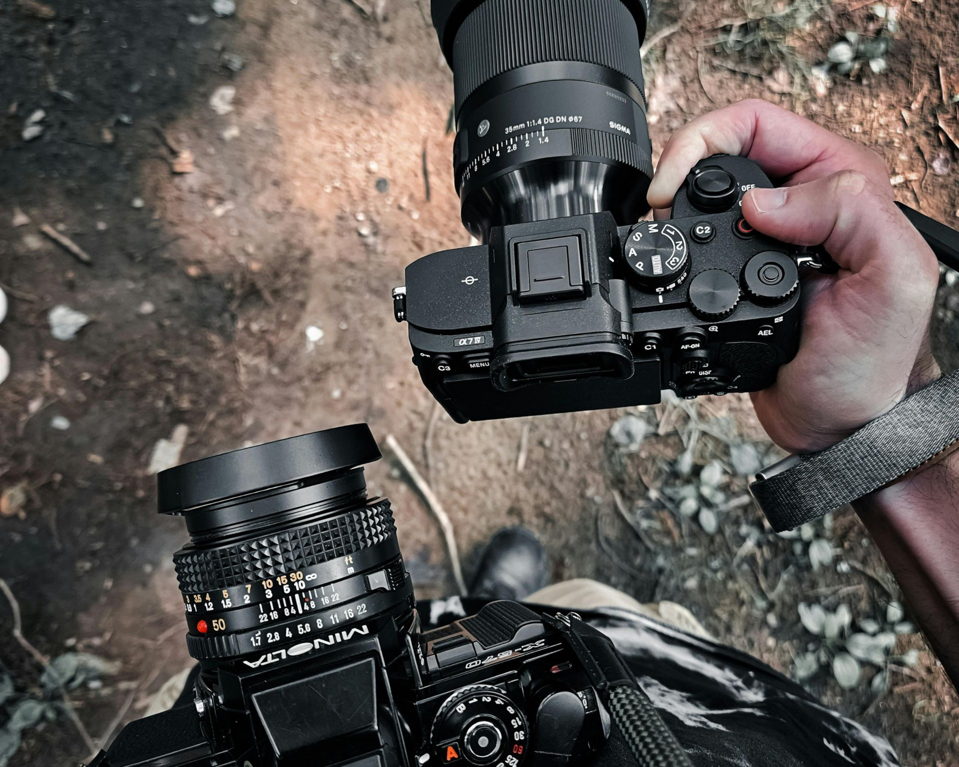 Two cameras held outdoors over rocky ground, one in each hand, with a lens cap and controls visible