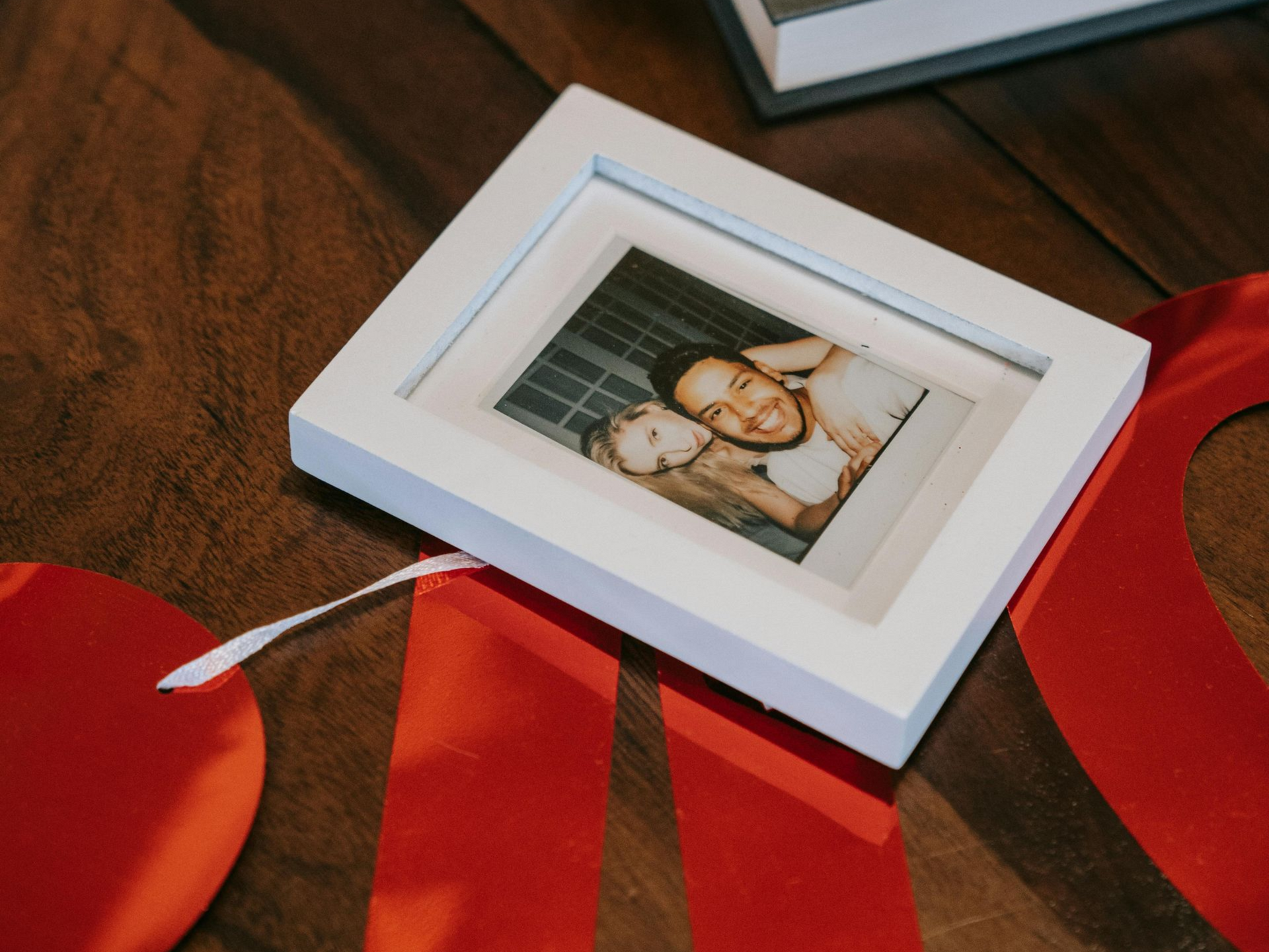 White photo frame with a smiling couple on a wooden table, surrounded by red ribbon and a red heart decoration