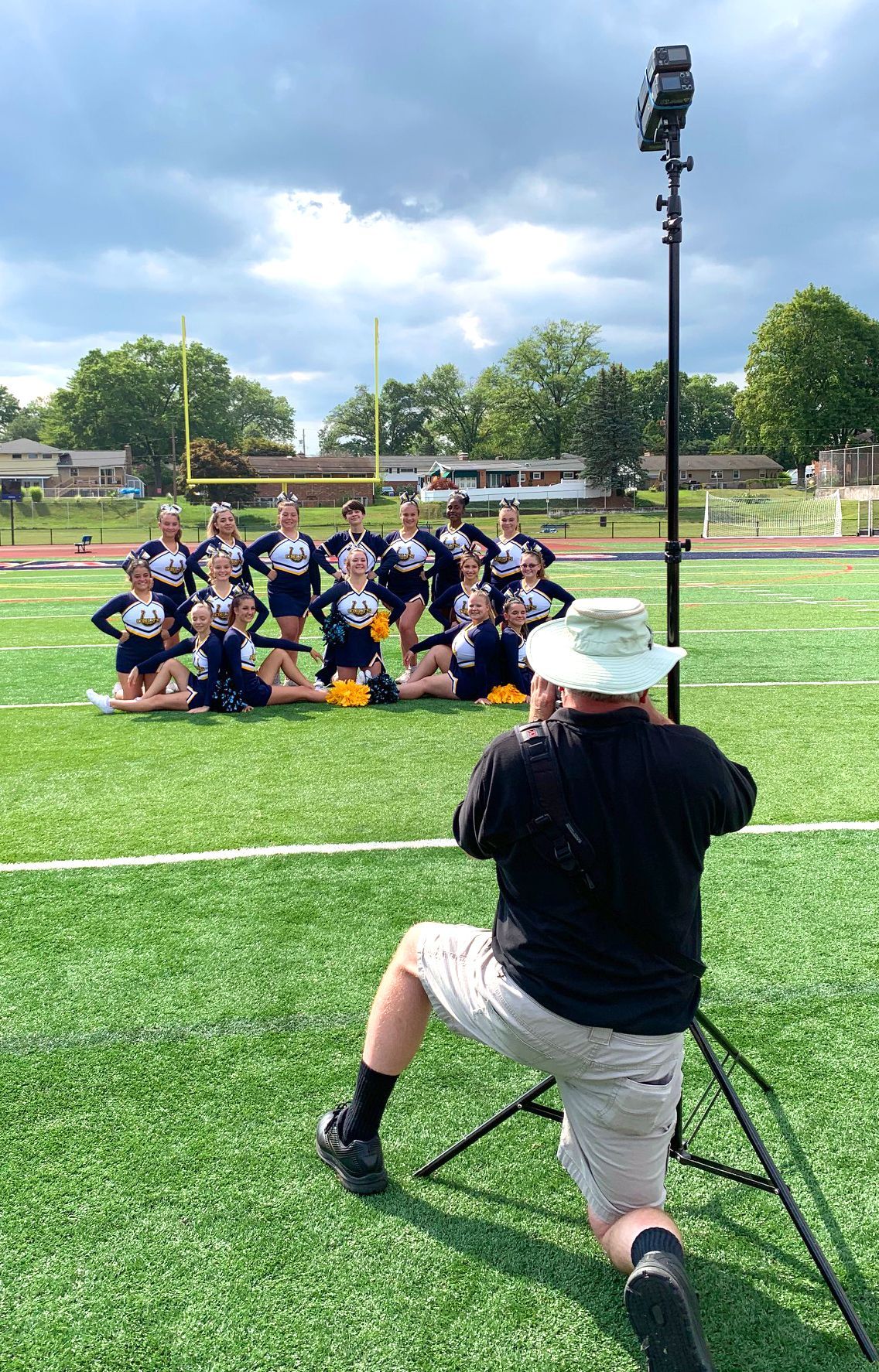 Photographer kneels on turf, aiming at a group posing on a sports field with trees and stadium lights behind them.