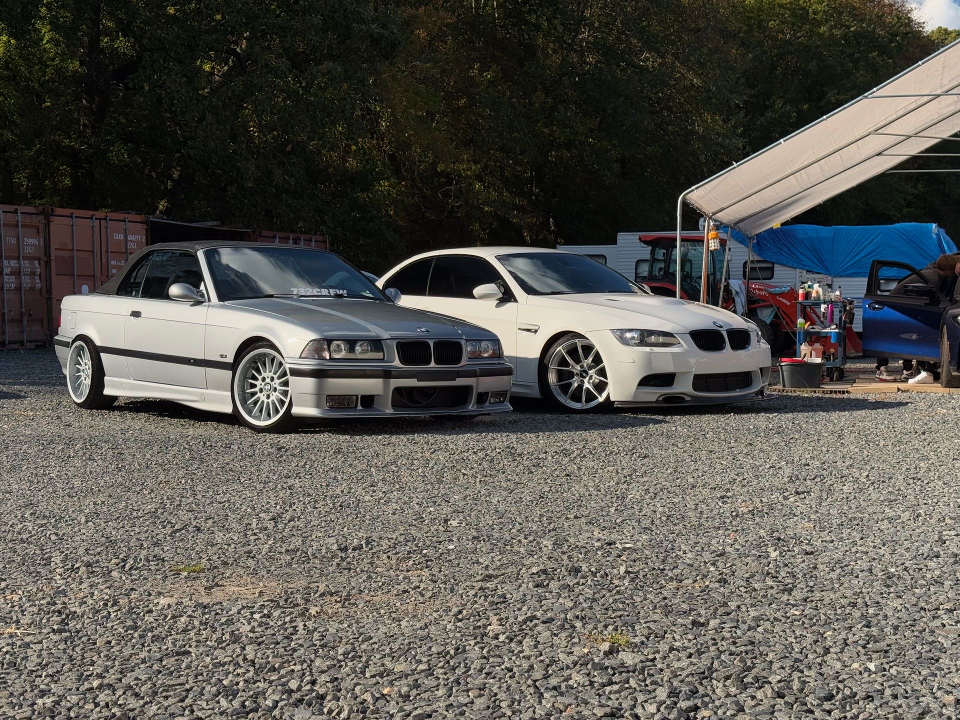 Two silver BMW sedans parked on gravel near tents and a canopy at an outdoor car meet