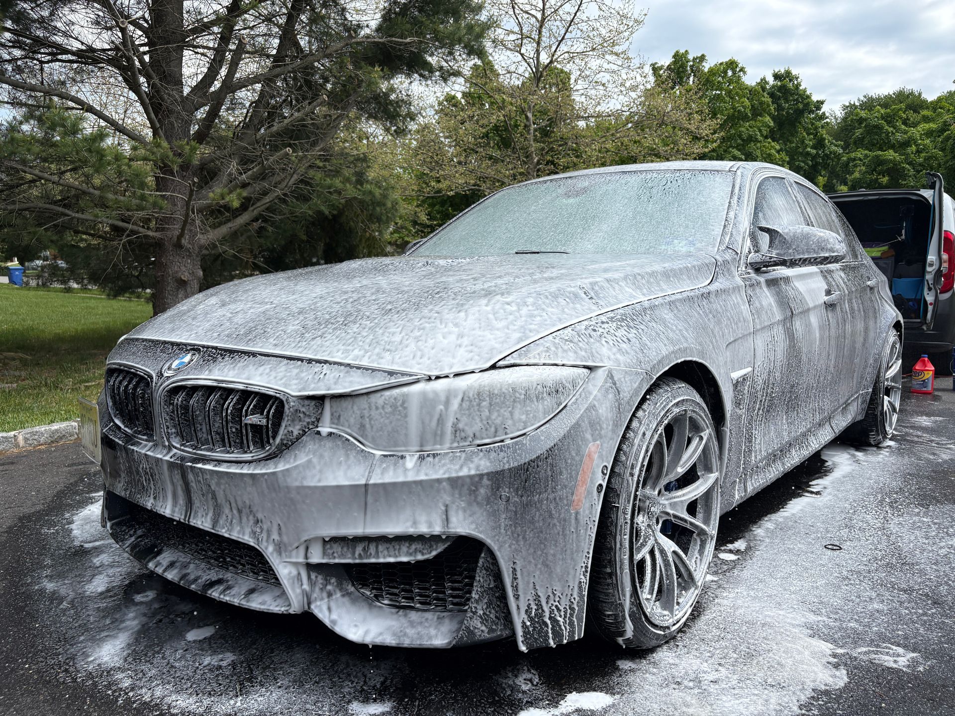 Silver BMW sedan covered in thick white foam during a car wash, parked outdoors on a wet driveway.
