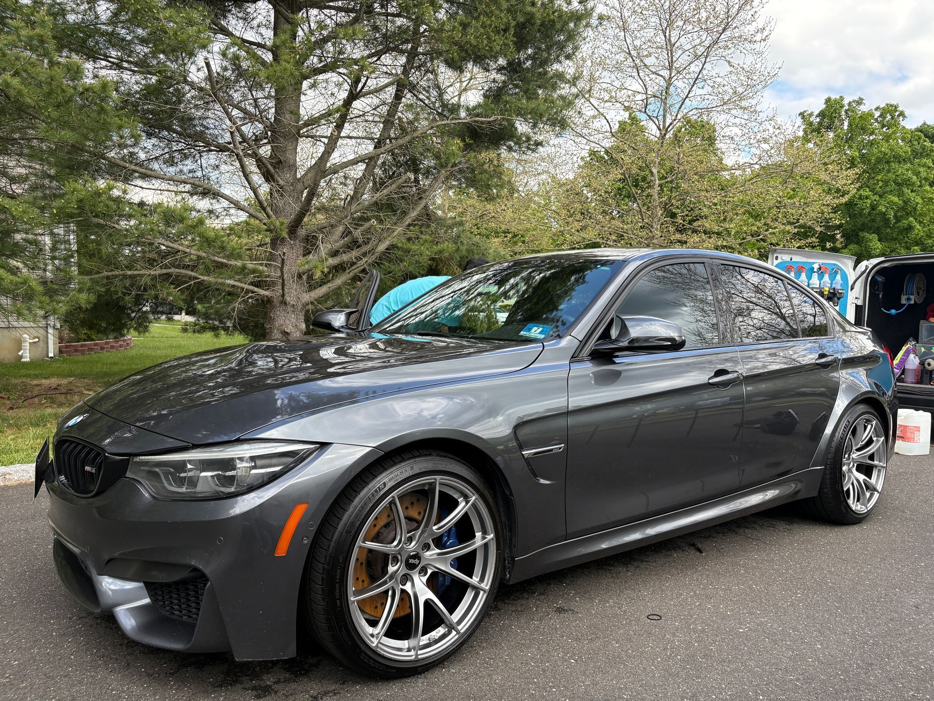 Dark gray BMW coupe parked outdoors on a wet road, with chrome wheels and trees in the background