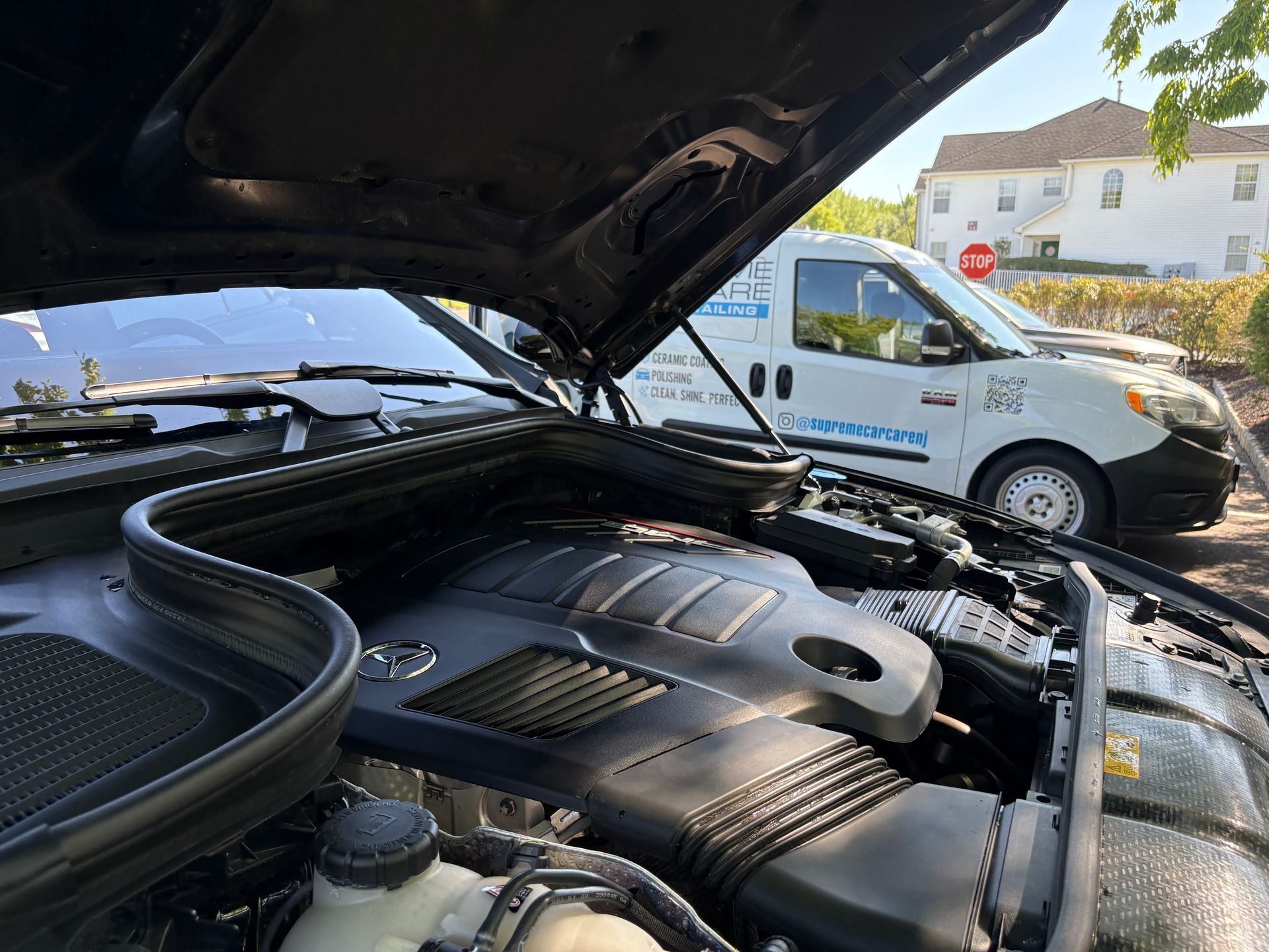 Open car hood showing engine bay, with a white service van parked beside it outdoors.
