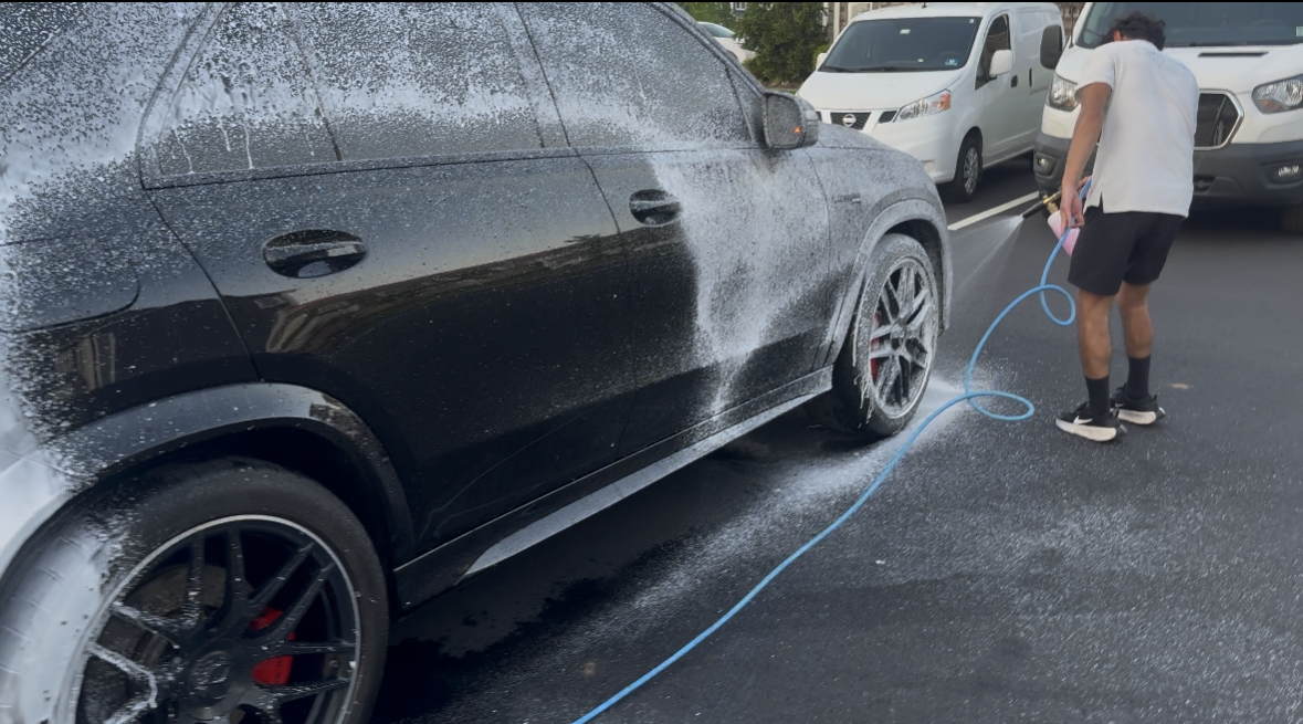 Person washing a black SUV with foam at a car wash, with a white van in the background