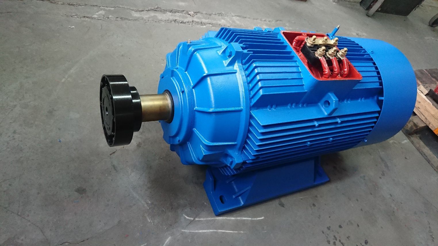 Motor eléctrico industrial azul sobre el suelo de un taller, con conexiones de terminales rojas en la parte superior.
