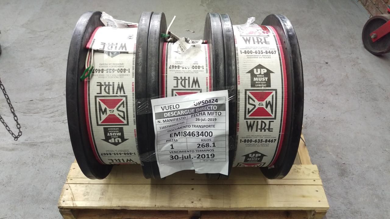 Tres grandes bobinas de alambre sobre un palé de madera, envueltas en etiquetas blancas con marcas rojas y negras.