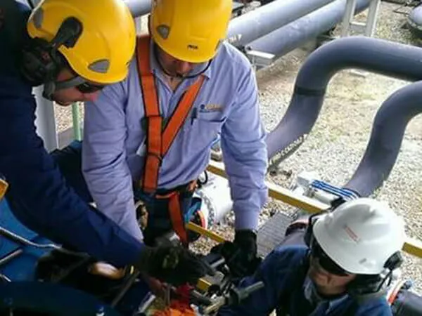 Tres trabajadores con cascos inspeccionan equipos en una plataforma de tuberías industriales.