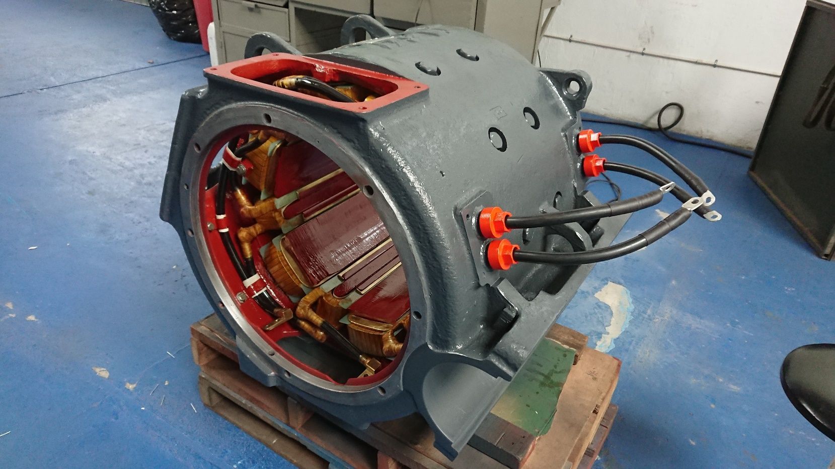 Motor industrial gris con bobinados de cobre expuestos y cableado rojo sobre un palé de madera en un taller.