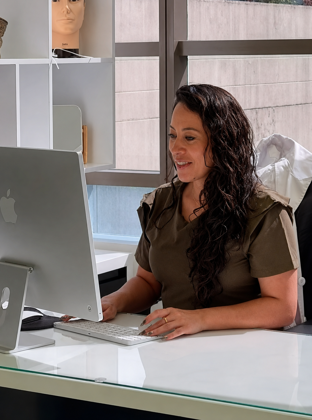 Dr Caballero smiling at a desk in a bright office, using a laptop beside a keyboard and monitor.