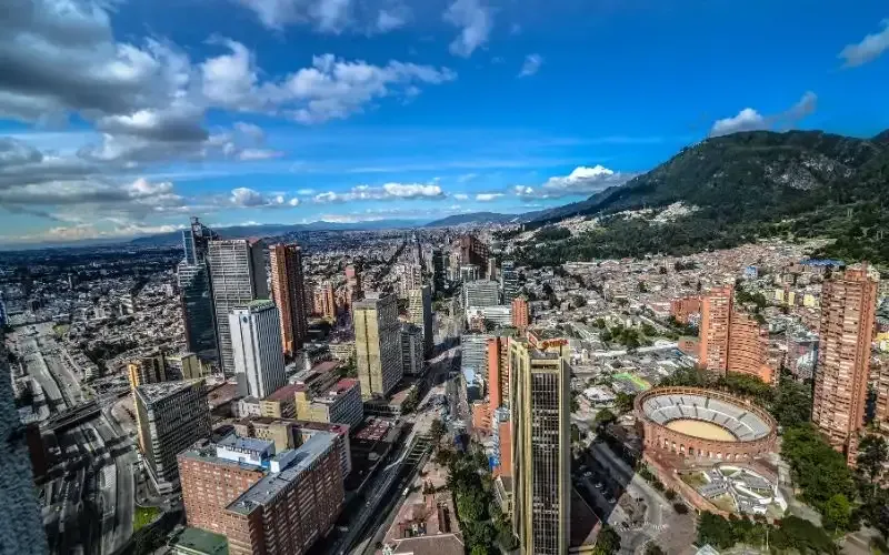 High-angle view of Bogotá, Colombia, with skyscrapers, city buildings, and a mountain in the background under a blue sky.