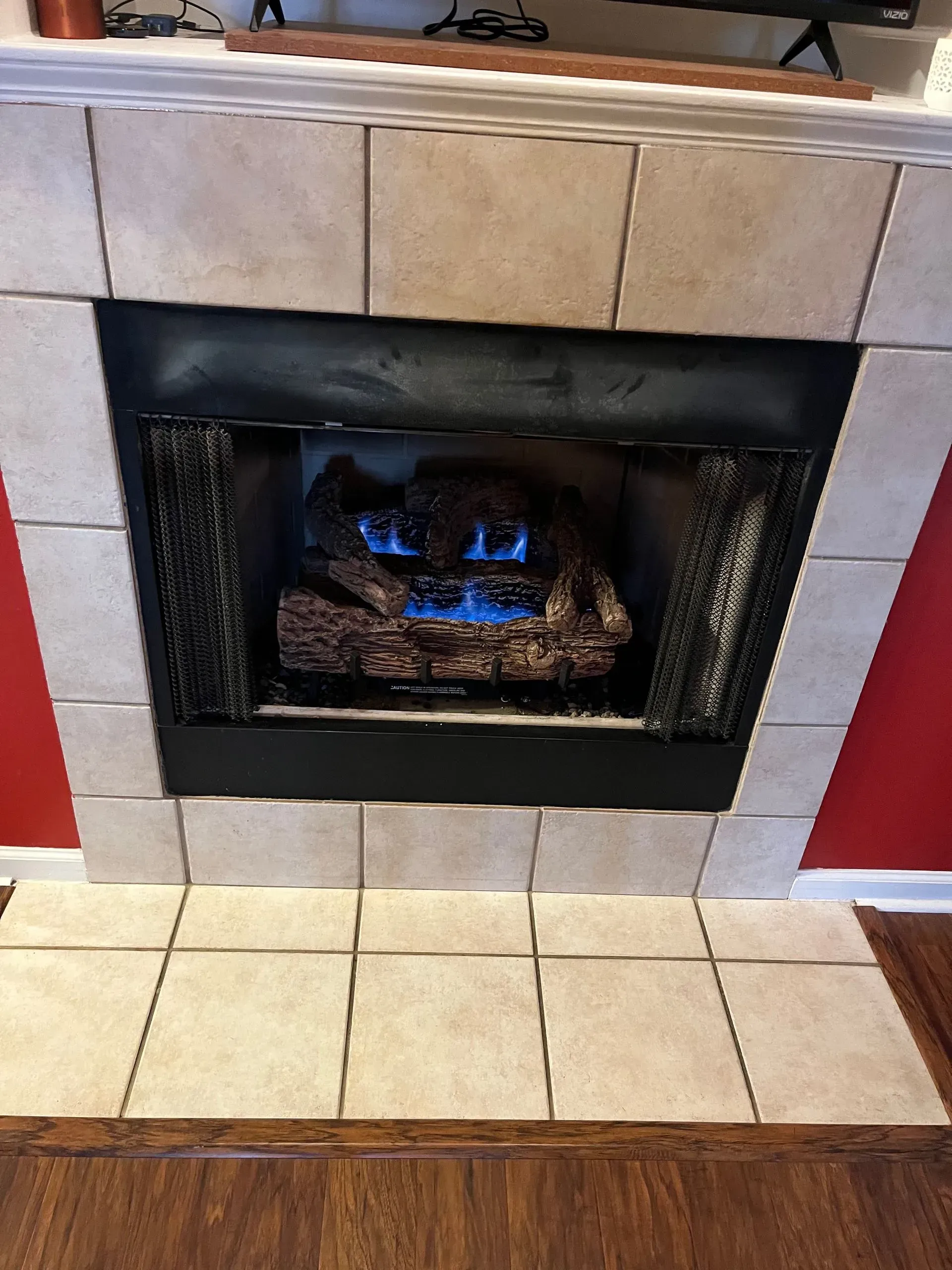 A fireplace with lit blue flames and faux logs, surrounded by cream-colored tiles, red wall, and wood flooring.