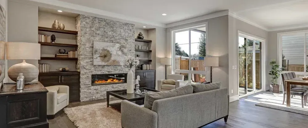 Living room with a stone fireplace, dark wood shelves, gray sofa, and large windows.