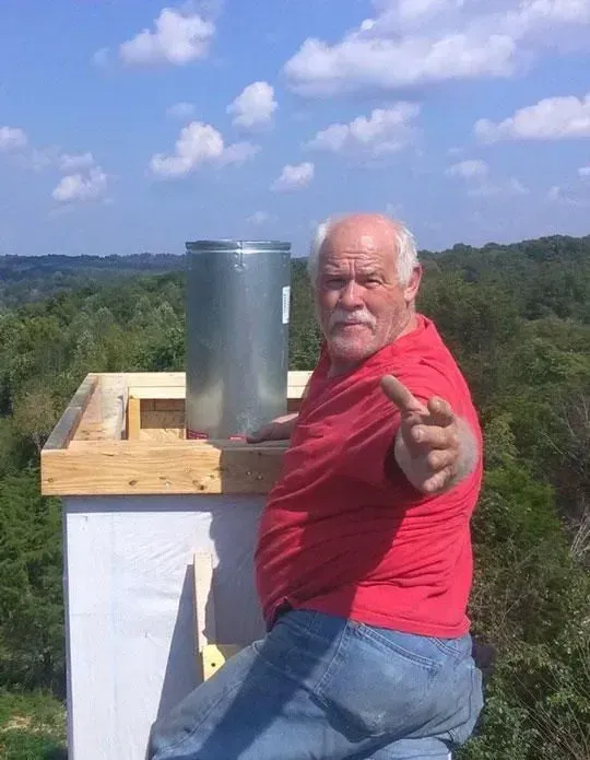 Man on roof pointing, next to metal chimney, wooded background. Red shirt, blue jeans, bright sky.