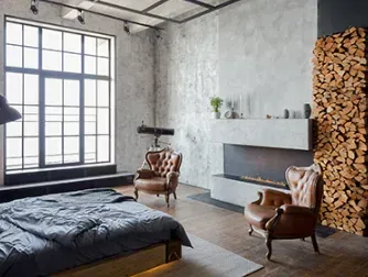 Bedroom with large window, fireplace, leather chairs, wood stack, and exposed concrete walls.