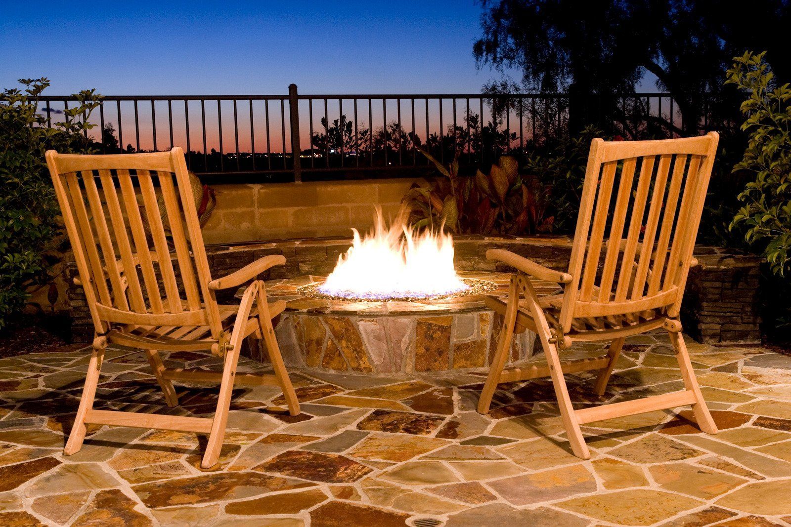 Two wooden chairs face a lit fire pit on a stone patio at dusk.