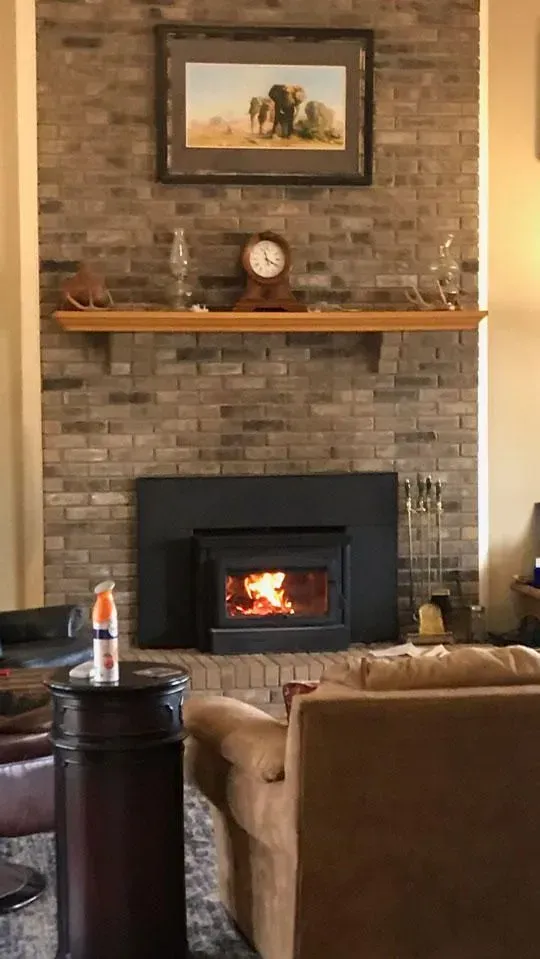 Fireplace with burning fire, brick backdrop, wooden mantel with decor. Brown couch and table in foreground.
