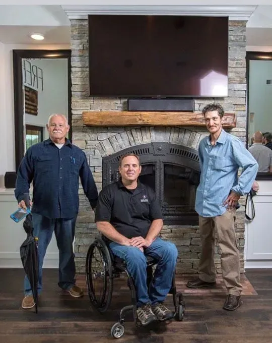 Three men near fireplace; one in wheelchair, others standing. Stone wall, TV, and mirror in background.
