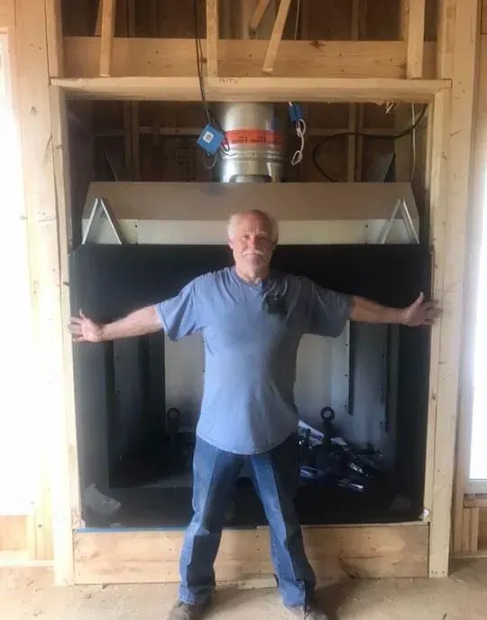 Man stands inside a fireplace opening, arms outstretched. The fireplace is framed in wood studs.