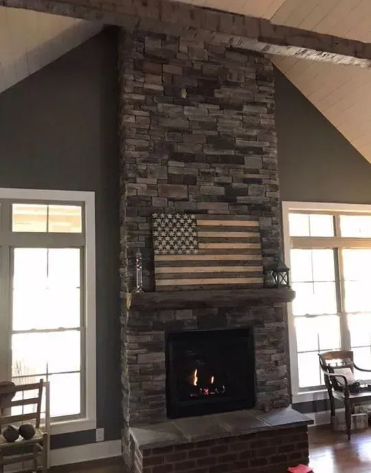 Stone fireplace with wooden flag, flanked by windows, inside a home.