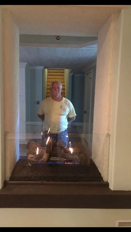 Man standing behind a modern fireplace with flames. White walls and hallway visible.