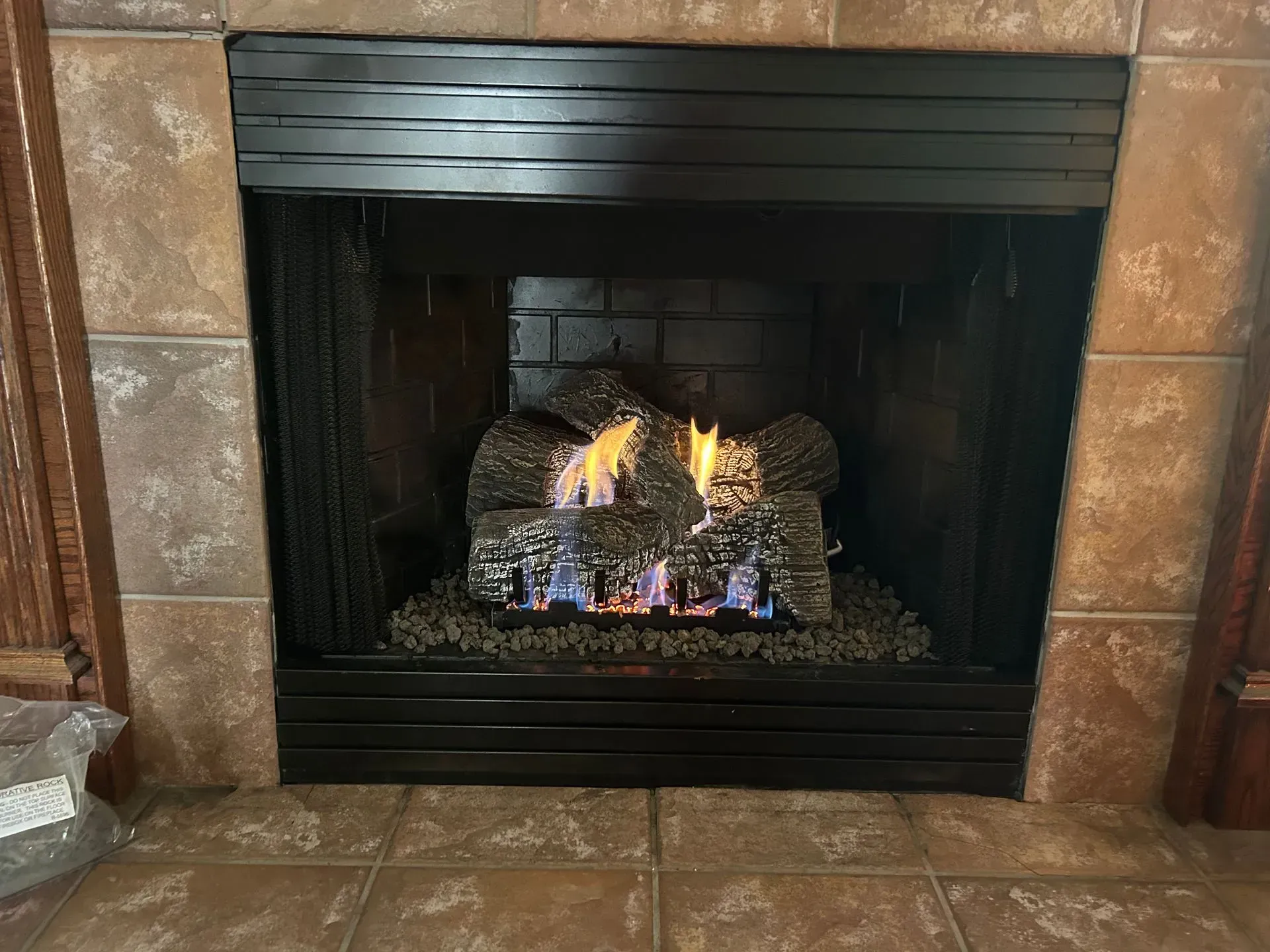 Gas fireplace with burning logs, inside a dark framed opening, surrounded by tile.
