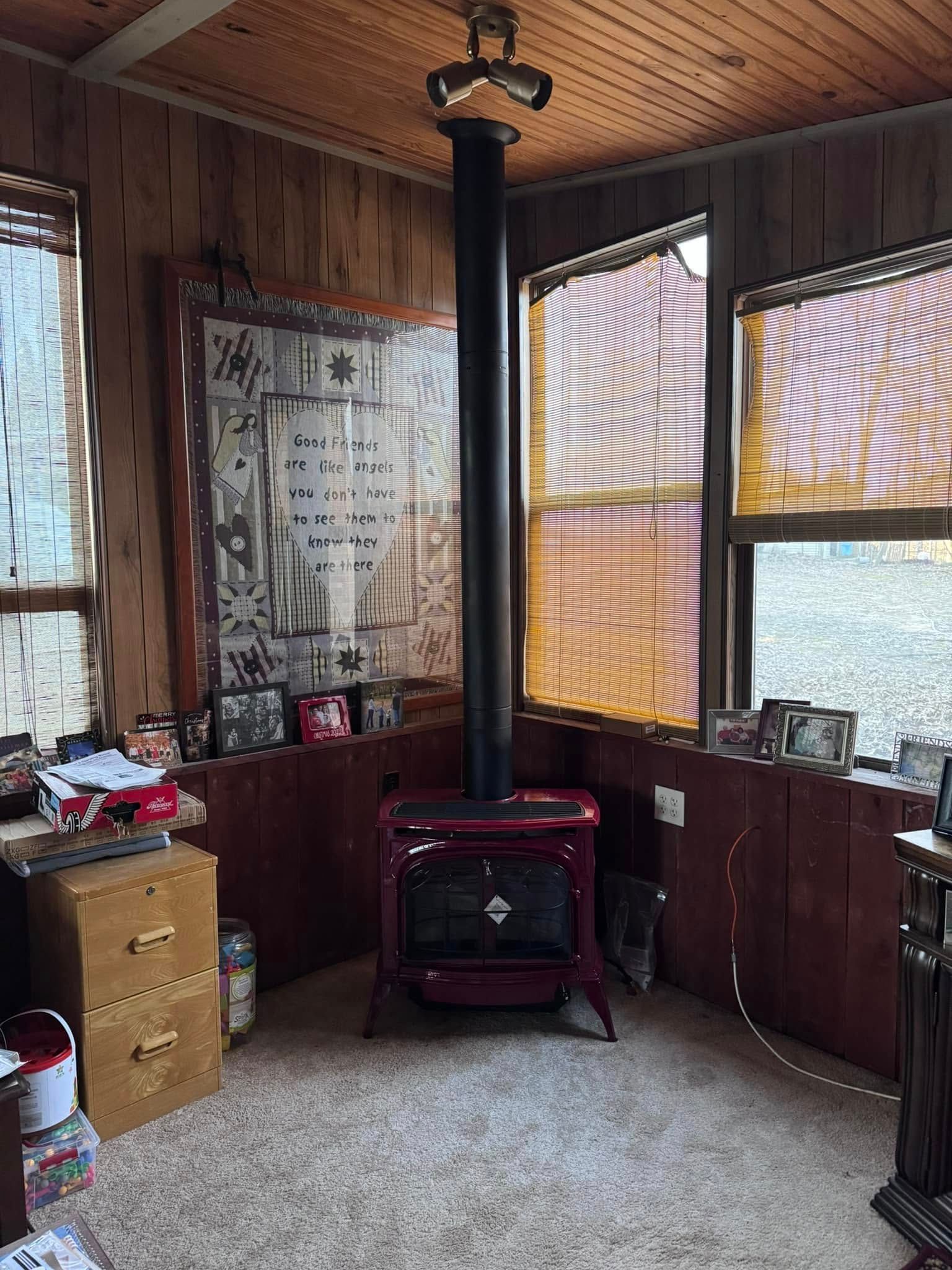 Red wood-burning stove with black chimney pipe, in corner room with wood paneling and blinds.