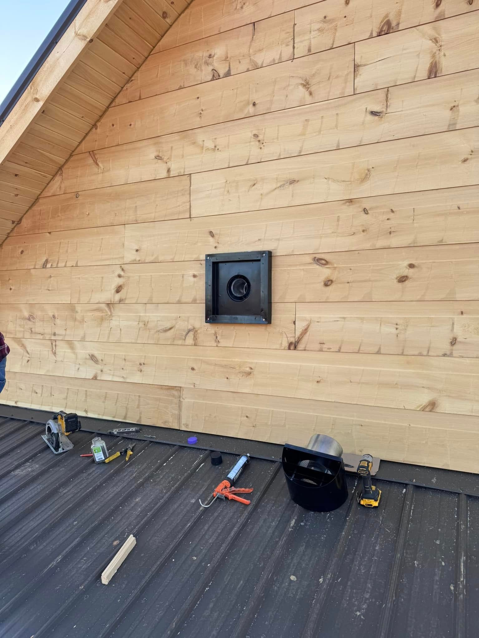 Black vent on wooden siding of a building with tools scattered on a metal roof.