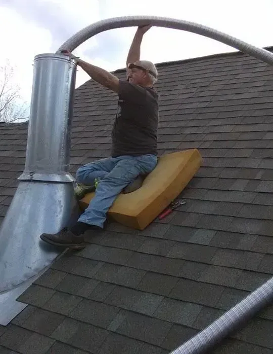 Man on a roof installing a flexible metal pipe onto a chimney. He sits on a foam pad.