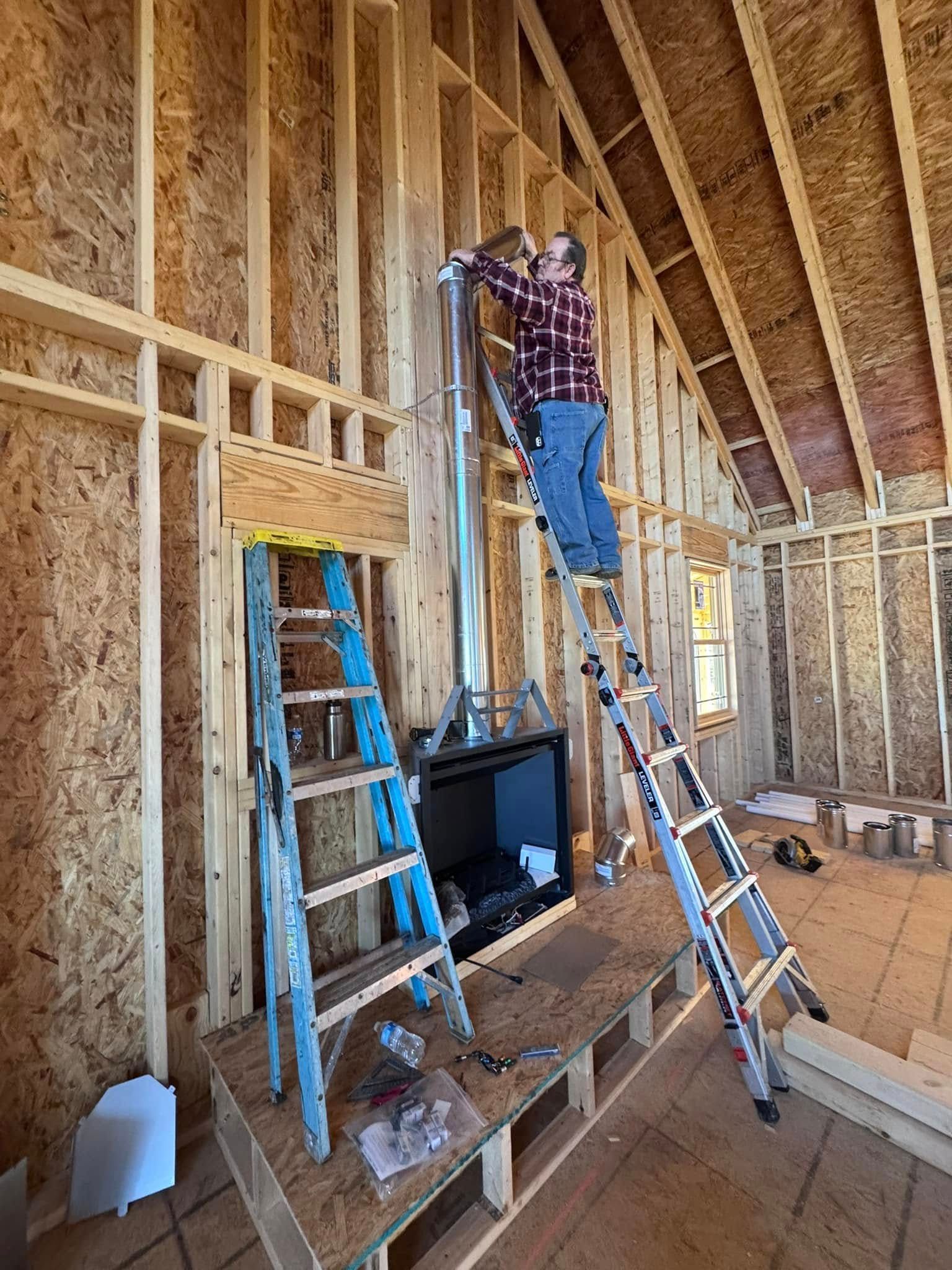 A person on a ladder installs a chimney flue in a partially built interior with wooden framing and a fireplace.