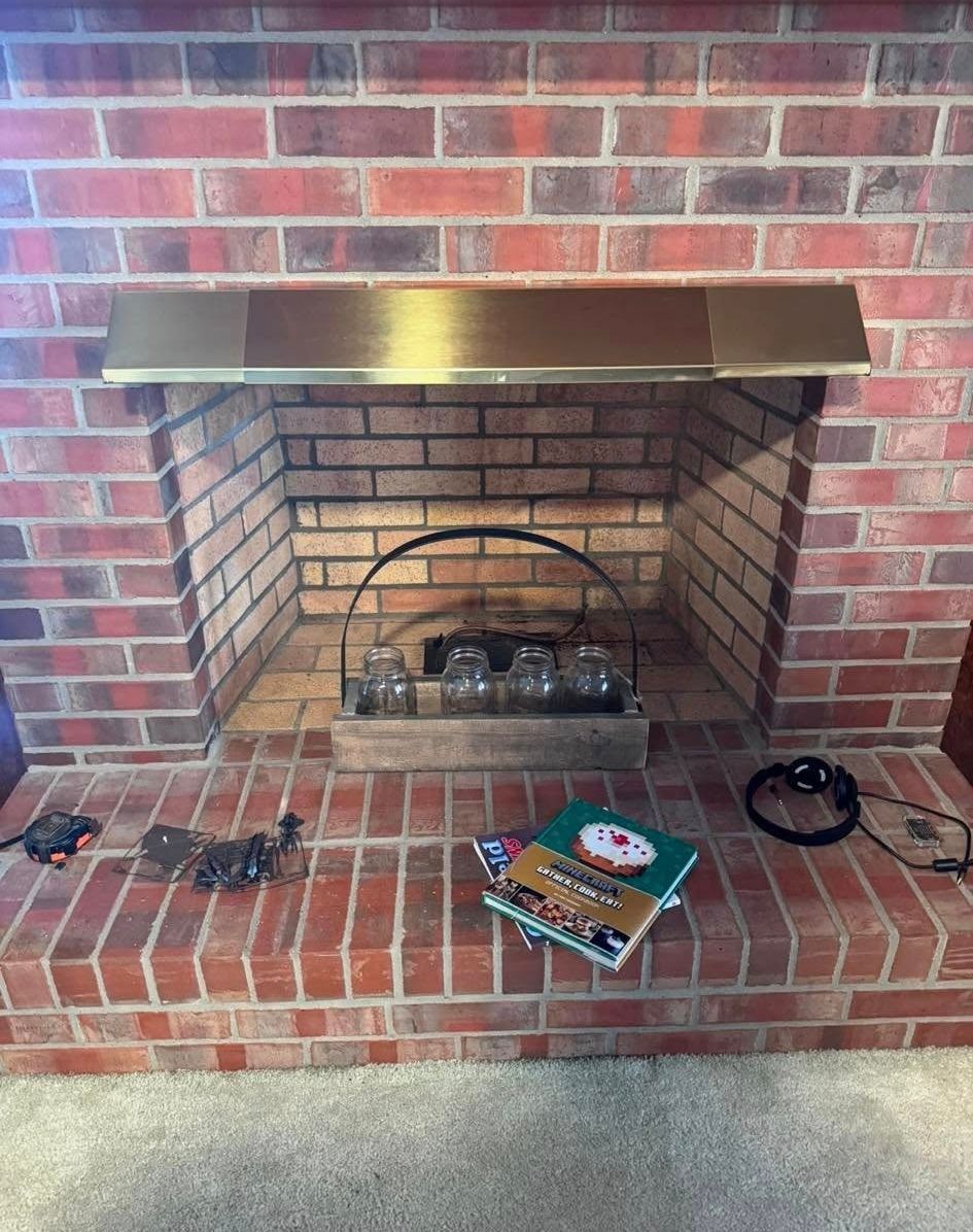 Brick fireplace with gold-colored metal trim and a black wire fire screen; books and electronics sit on the hearth.