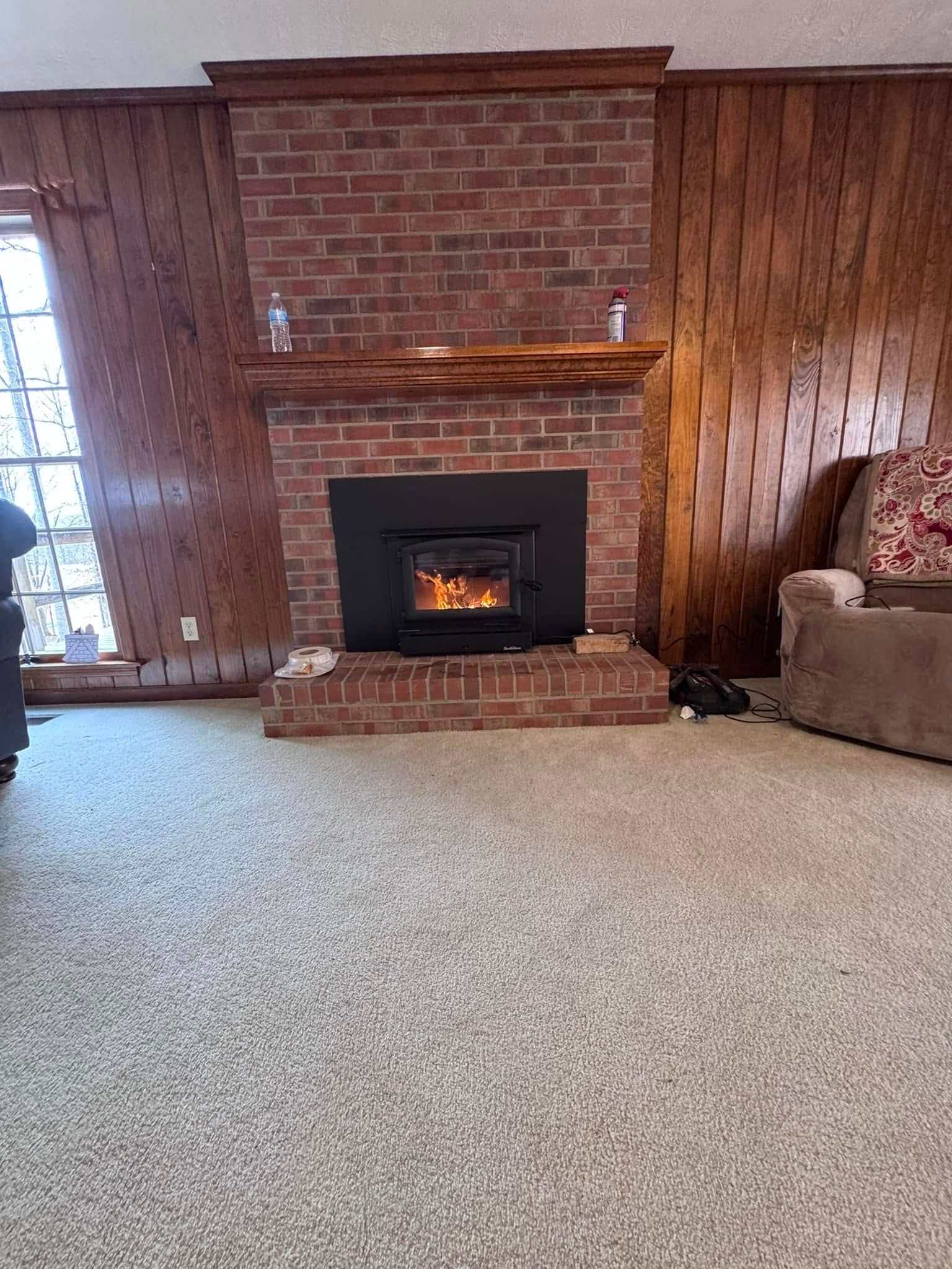 A brick fireplace with a burning fire, flanked by wood-paneled walls and a carpeted floor.