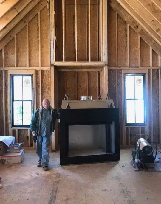 Man stands near unfinished fireplace in a wooden-framed room with windows. Construction setting.