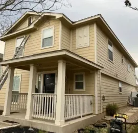 A two-story yellow house under renovation with workers, ladders, and painting equipment on the exterior.