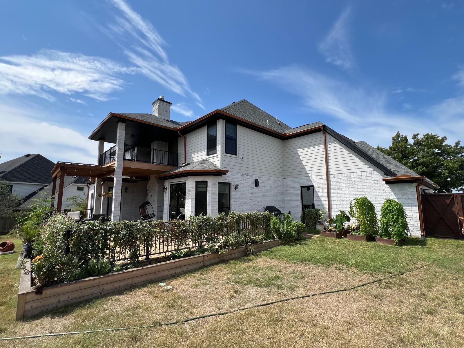 A two-story white brick house with a dark roof and a wooden deck on a sunny day with a clear blue sky.