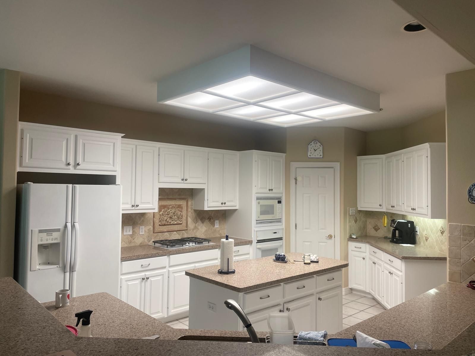 A white-cabinet kitchen with beige countertops, a central island, stainless appliances, and a large grid ceiling light.