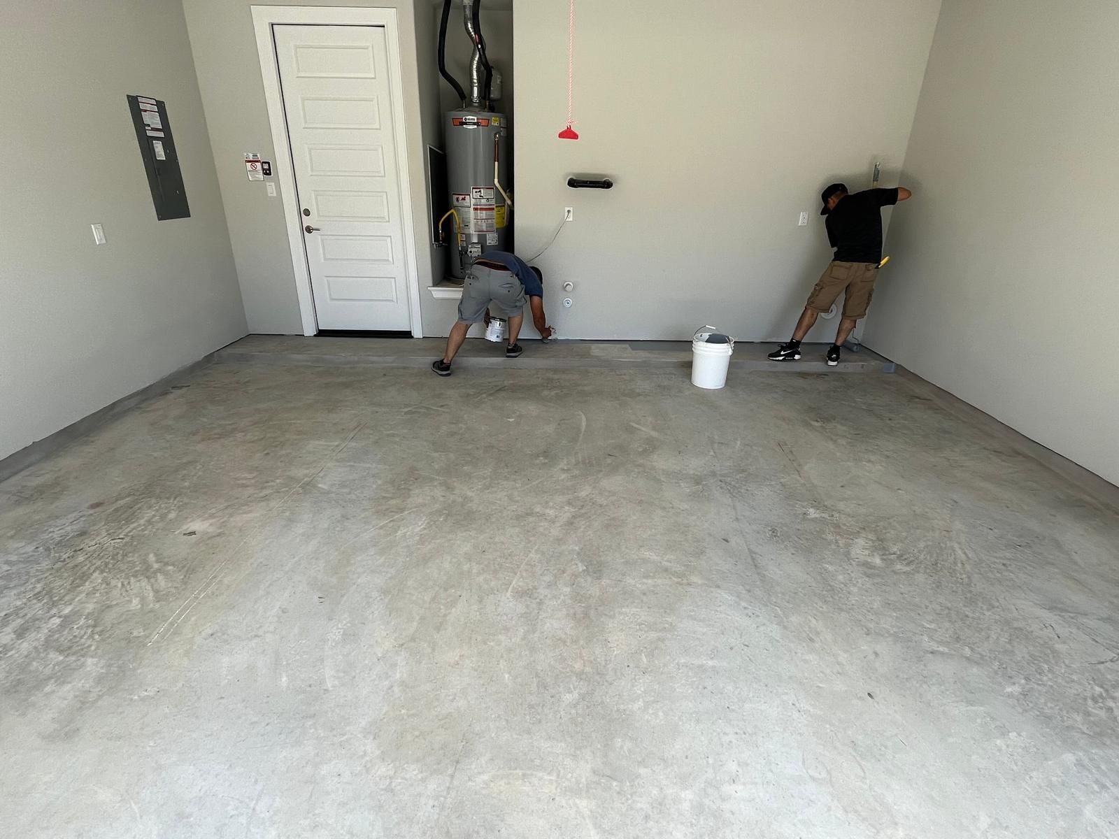 Two workers prepare a concrete garage floor for coating, with one near a water heater and one near a corner.