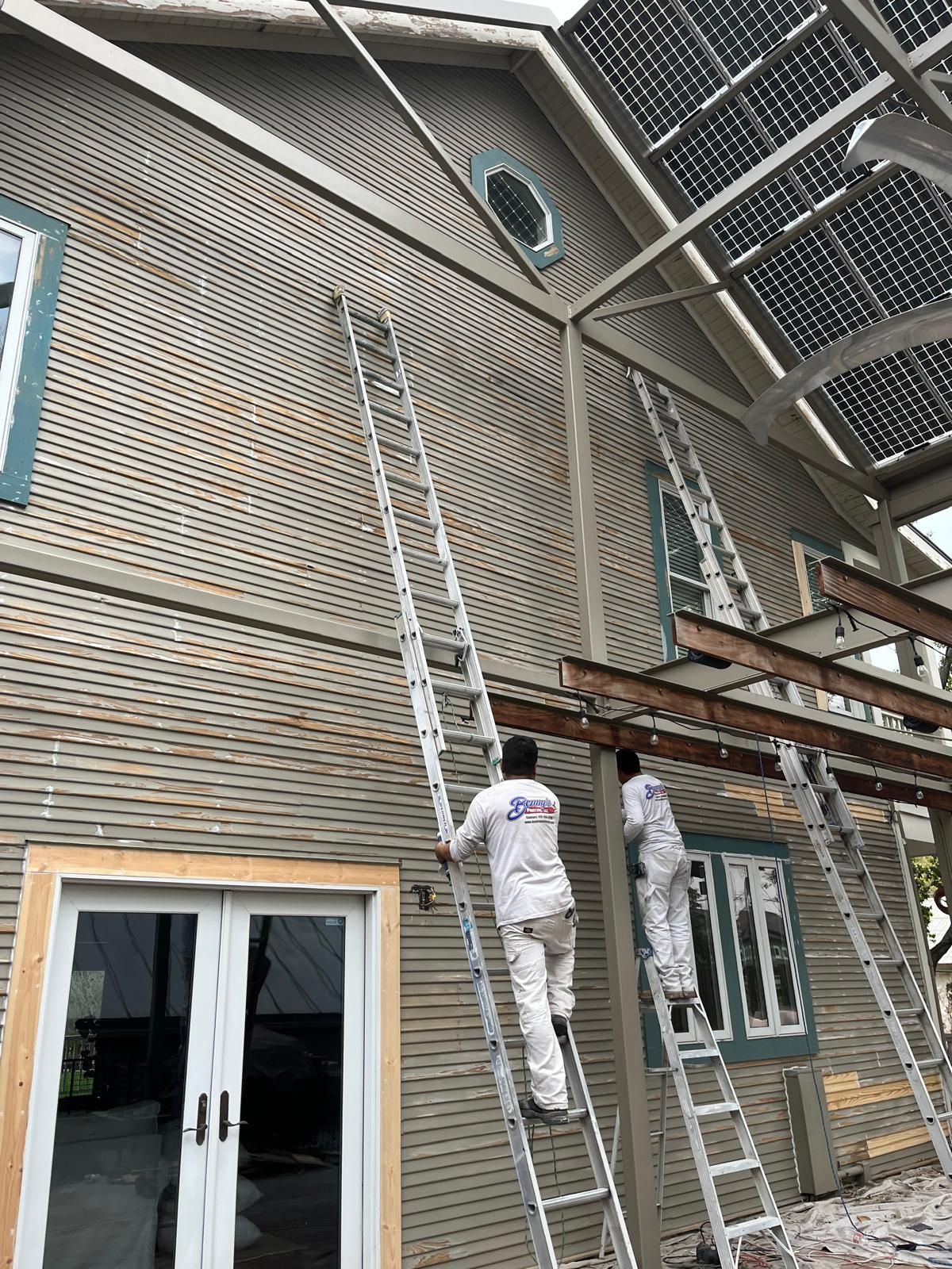 Two workers in white uniforms climb extension ladders placed against the gray-sided wall of a two-story house.