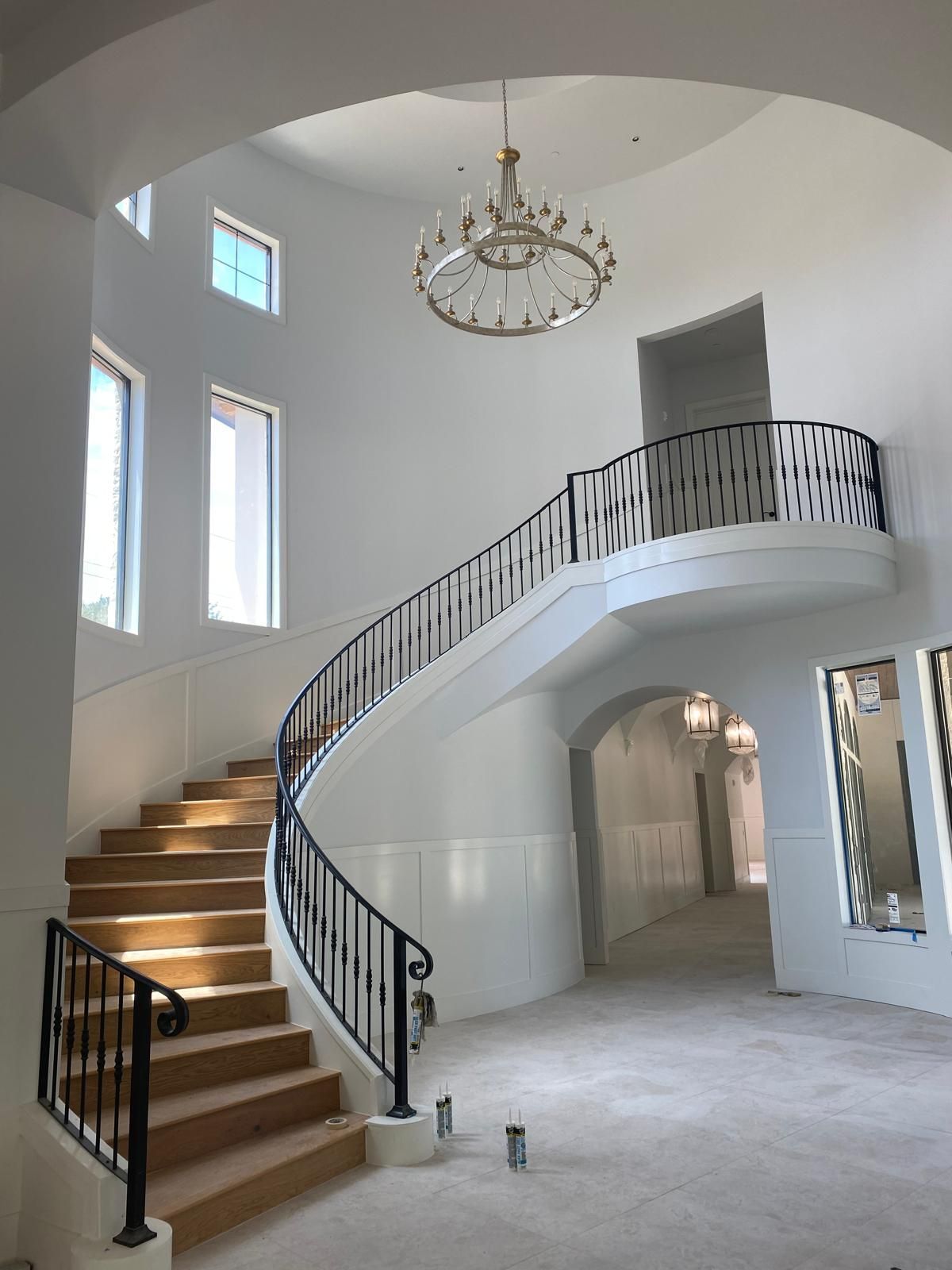 A curved staircase with black iron railings leads to a landing in a large, white-walled foyer with a hanging chandelier.