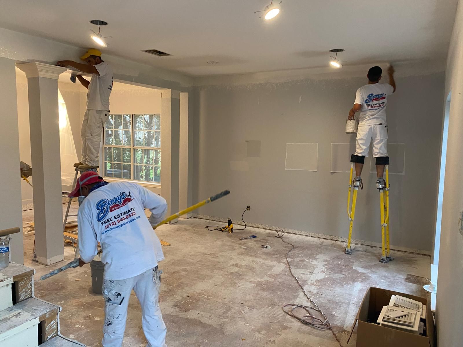 Three construction workers in uniform paint the walls and ceiling of a room, with one using stilts to reach high areas.