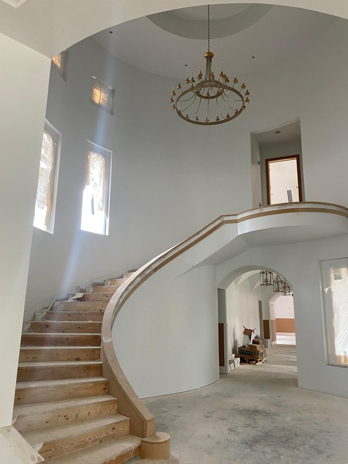 A grand, unfinished foyer with a curved wooden staircase, light-colored walls, and a large chandelier hanging from above.
